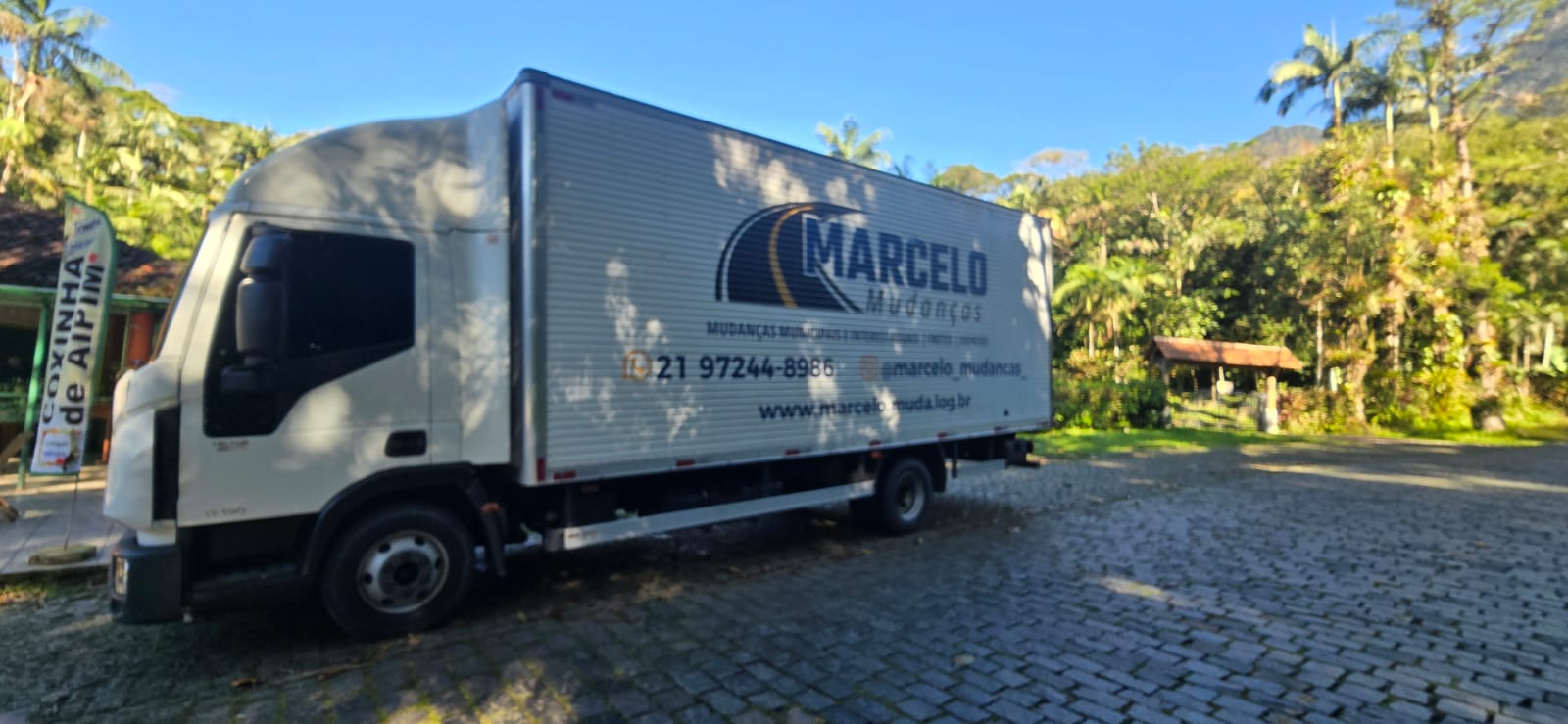 Baú da Marcelo Mudanças parado em estrada de pedra no meio da mata, com a logo e o telefone na lateral